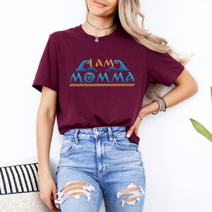 May include: A maroon t-shirt with the text "I AM MOMMA" in blue and gold lettering. The text is stylized with a wave design and a spiral.