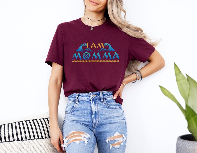 May include: A maroon t-shirt with the text "I AM MOMMA" in blue and gold lettering. The text is stylized with a wave design and a spiral.