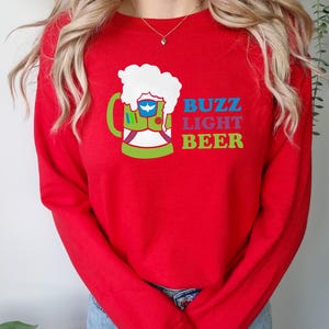 May include: Red long-sleeved sweatshirt featuring a graphic of a green beer mug with a white foam top. The mug contains a blue and white space ranger. The text "Buzz Light Beer" is printed below the mug in rainbow colours.