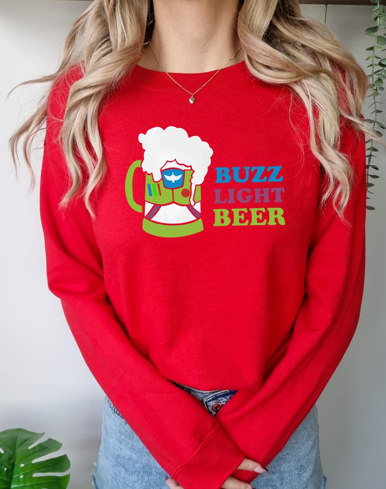 May include: Red long-sleeved sweatshirt featuring a graphic of a green beer mug with a white foam top. The mug contains a blue and white space ranger. The text "Buzz Light Beer" is printed below the mug in rainbow colours.