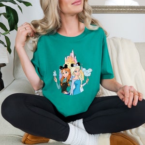 Op de afbeelding: Een turquoise t-shirt met een grafische print van twee Disney-prinsessen, Anna en Elsa, die Mickey Mouse-oren dragen en snacks vasthouden voor een kleurrijk kasteel.