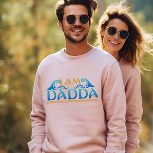 May include: A pink sweatshirt with a blue and yellow graphic that says "I AM DADDA" with waves and a gold border.