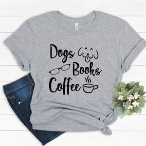 May include: A heather gray short-sleeve t-shirt featuring black text that reads Dogs, Books, and Coffee. The design includes a line-art illustration of a dog face, a pair of eyeglasses, and a steaming coffee cup. The shirt is knotted at the side.