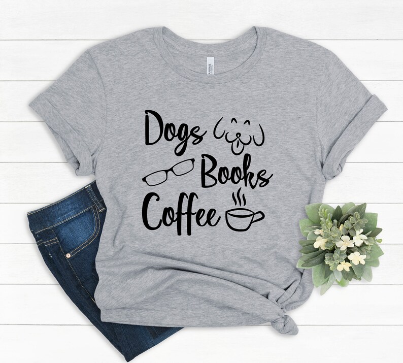 May include: A heather gray short-sleeve t-shirt featuring black text that reads Dogs, Books, and Coffee. The design includes a line-art illustration of a dog face, a pair of eyeglasses, and a steaming coffee cup. The shirt is knotted at the side.