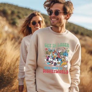 Puede incluir: Una sudadera beige con un estampado gr&aacute;fico de Mickey Mouse y otros personajes de Disney. El texto en la sudadera dice "Get in loser, we're going to Disney World."