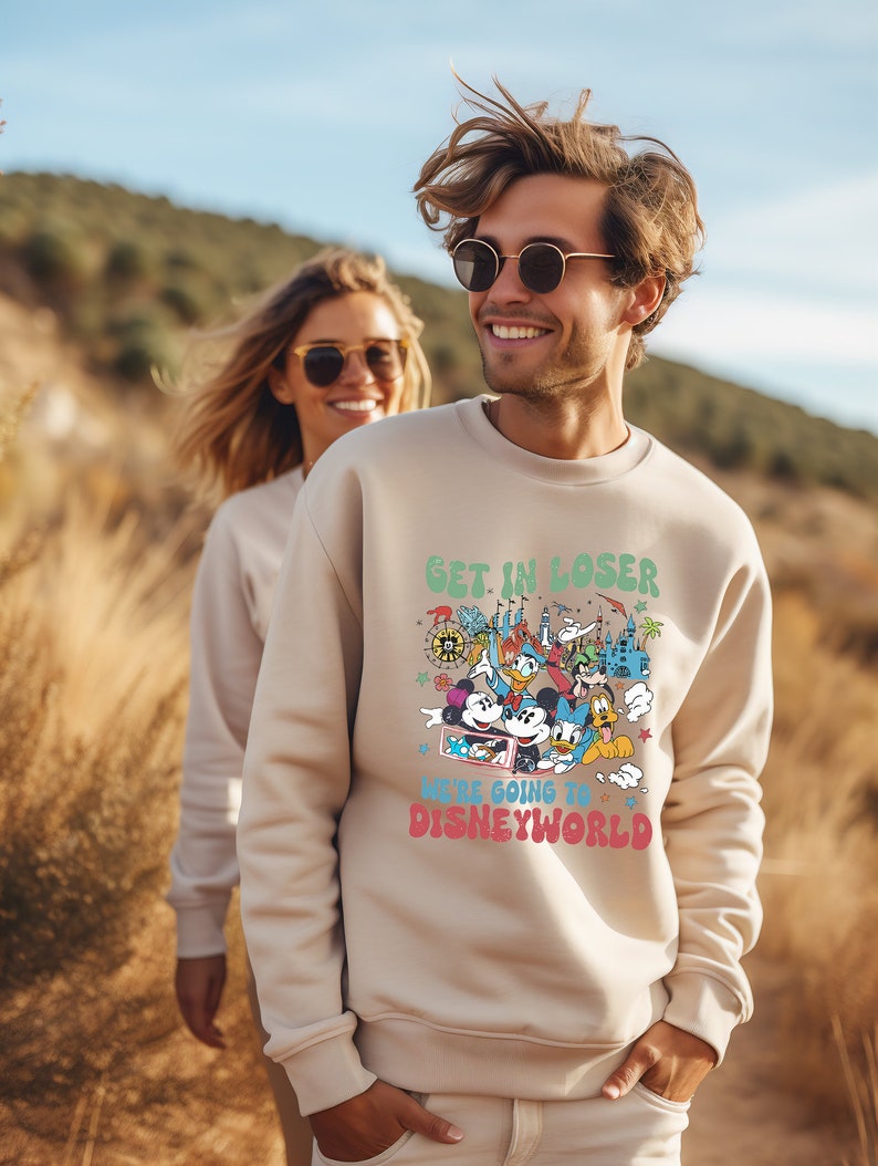 Puede incluir: Una sudadera beige con un estampado gr&aacute;fico de Mickey Mouse y otros personajes de Disney. El texto en la sudadera dice "Get in loser, we're going to Disney World."