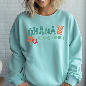 May include: A pale blue sweatshirt featuring the phrase "OHANA MEANS FAMILY" in green and orange, with a tiki mask and floral accents. The top is a crew neck style, suitable for casual wear.