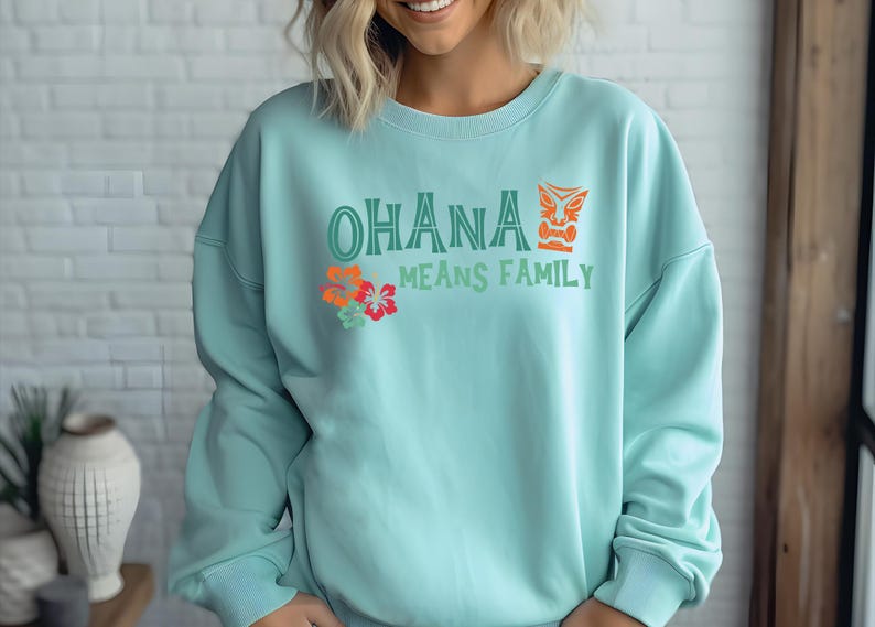 May include: A pale blue sweatshirt featuring the phrase "OHANA MEANS FAMILY" in green and orange, with a tiki mask and floral accents. The top is a crew neck style, suitable for casual wear.
