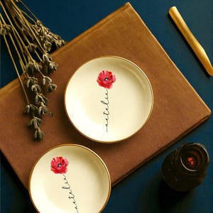 May include: Two small, off-white ceramic plates with gold rims, each featuring a red poppy and the word "Natalie" in cursive. The plates rest on a brown surface, with a gold-colored stick and a dark brown object nearby.