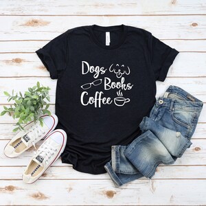 May include: A black short-sleeved t-shirt featuring white text that reads Dogs, Books, and Coffee, accompanied by graphic icons of a dog face, eyeglasses, and a steaming coffee cup, paired with blue denim jeans and white canvas sneakers.