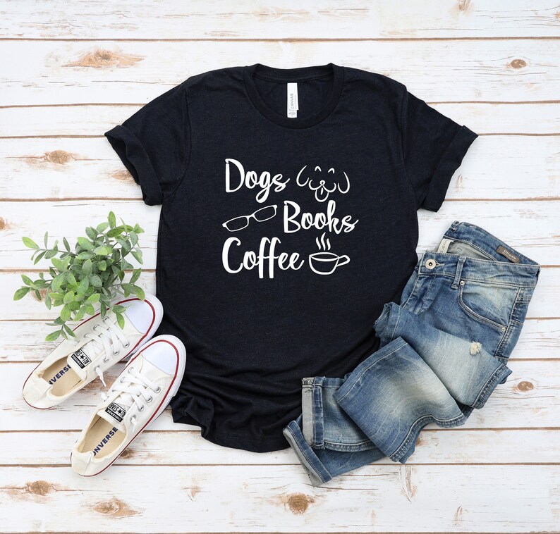 May include: A black short-sleeved t-shirt featuring white text that reads Dogs, Books, and Coffee, accompanied by graphic icons of a dog face, eyeglasses, and a steaming coffee cup, paired with blue denim jeans and white canvas sneakers.