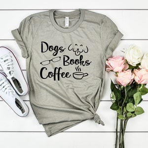 May include: A heather gray short-sleeve t-shirt featuring black text that reads Dogs, Books, and Coffee, accompanied by graphic icons of a dog face, eyeglasses, and a steaming coffee cup, paired with white sneakers and a bouquet of pink and white roses.