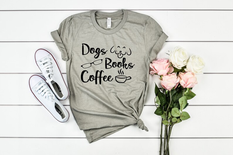 May include: A heather gray short-sleeve t-shirt featuring black text that reads Dogs, Books, and Coffee, accompanied by graphic icons of a dog face, eyeglasses, and a steaming coffee cup, paired with white sneakers and a bouquet of pink and white roses.