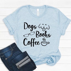 May include: A light blue short-sleeve t-shirt featuring black text that reads Dogs, Books, and Coffee. The design includes a black graphic of a dog face, a pair of eyeglasses, and a steaming coffee cup. Dark blue denim jeans are positioned below.
