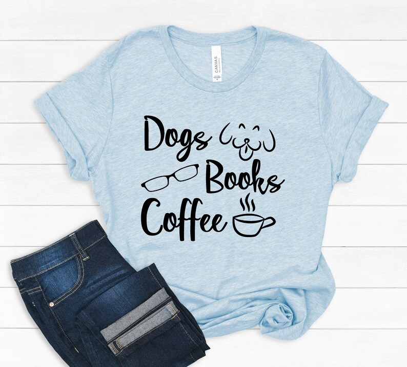 May include: A light blue short-sleeve t-shirt featuring black text that reads Dogs, Books, and Coffee. The design includes a black graphic of a dog face, a pair of eyeglasses, and a steaming coffee cup. Dark blue denim jeans are positioned below.