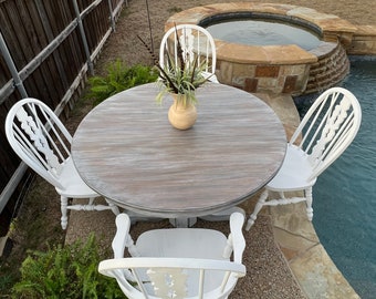 May include: A round wooden dining table with a gray-washed finish, surrounded by four white chairs. A beige vase with greenery sits in the center. The table and chairs are set up outdoors near a pool.