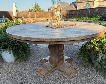 May include: A large, round wooden outdoor dining table with a pedestal base. The table is topped with a clear glass vase filled with decorative balls and greenery. The table is surrounded by greenery and is set on a concrete patio.