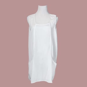 May include: A sleeveless white dress with a square neckline and two front pockets. The dress is displayed on a white mannequin against a muted pink background. The fabric appears to be lightweight.