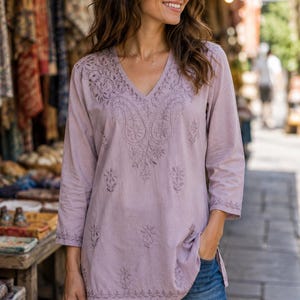 May include: A light purple tunic featuring a V-neckline and three-quarter length sleeves. The garment is decorated with tonal floral and paisley embroidery patterns across the chest, sleeves, and hemline, paired with blue denim jeans.