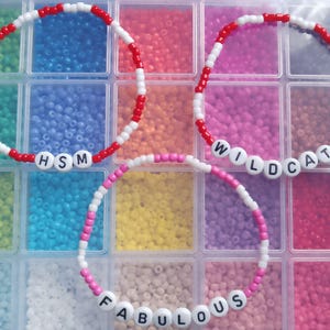 Peut inclure: Trois bracelets de perles avec des perles de lettres blanches épelant "HSM", "WILDCATS" et "FABULOUS". Les bracelets sont faits de perles rouges, blanches et roses. Les bracelets sont exposés dans un organisateur en plastique transparent rempli de perles colorées.