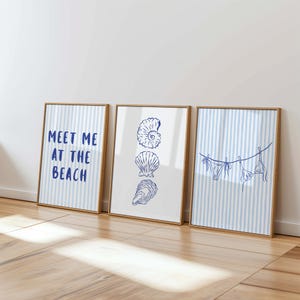 Könnte beinhalten: Drei gerahmte Kunstdrucke mit Strandmotiven. Ein Druck zeigt den Text "MEET ME AT THE BEACH" in Blau. Die anderen Drucke zeigen blaue Strichzeichnungen von Muscheln und einem Bikini auf einer Wäscheleine. Die Rahmen sind hellbraun.