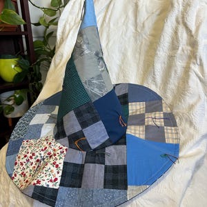 May include: A pointed hat constructed from a patchwork of various blue, gray, and white fabric squares. The design features a mix of solid colors, plaid patterns, and a floral print section, with small colorful thread ties visible on the brim.