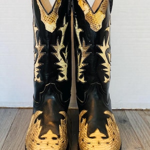 May include: A pair of cowboy boots with a black leather upper and a snakeskin-patterned vamp and shaft. The boots have a pointed toe and decorative stitching. The shaft features a decorative cutout design. The boots are set against a light background.