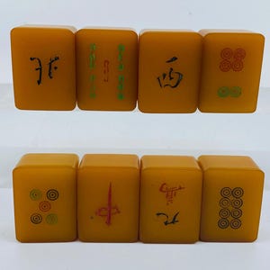 Op de afbeelding: Acht vintage, amberkleurige Mahjong-tegels. Elke tegel heeft zwarte of groene symbolen en karakters. De tegels zijn gerangschikt in twee rijen van vier, die de verscheidenheid aan ontwerpen laten zien.