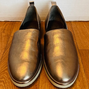 May include: A pair of metallic gold slip-on shoes with a white rubber sole.