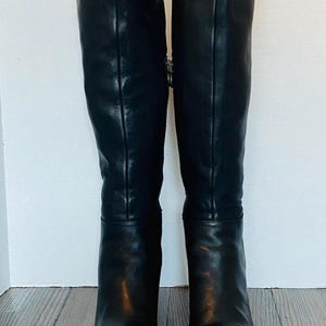 May include: A pair of black knee-high boots featuring a smooth leather-like finish, a rounded toe, and a vertical seam running up the front of the shaft. The boots have a side zipper closure and a block heel, providing a structured and tall silhouette.
