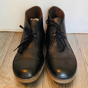 May include: A pair of black leather chukka boots with black laces. The boots have a light brown sole and a dark brown interior. The shoes are displayed on a light brown wooden surface.