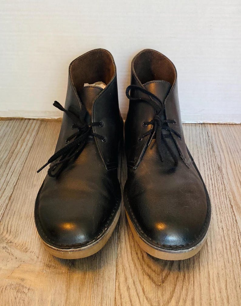 May include: A pair of black leather chukka boots with black laces. The boots have a light brown sole and a dark brown interior. The shoes are displayed on a light brown wooden surface.