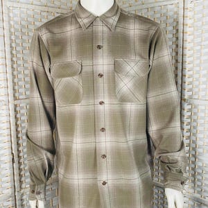 May include: A long-sleeved, button-down shirt in a plaid pattern. The shirt features two chest pockets and a collared neckline. The color palette includes shades of olive green, beige, and white. The shirt is displayed on a mannequin.