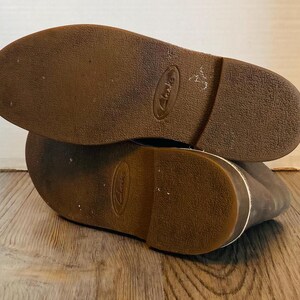May include: Close-up of the brown leather soles of a pair of brown leather shoes. The shoes have a white stitching detail around the edges of the soles. The brand name "Clarks" is visible on the soles.