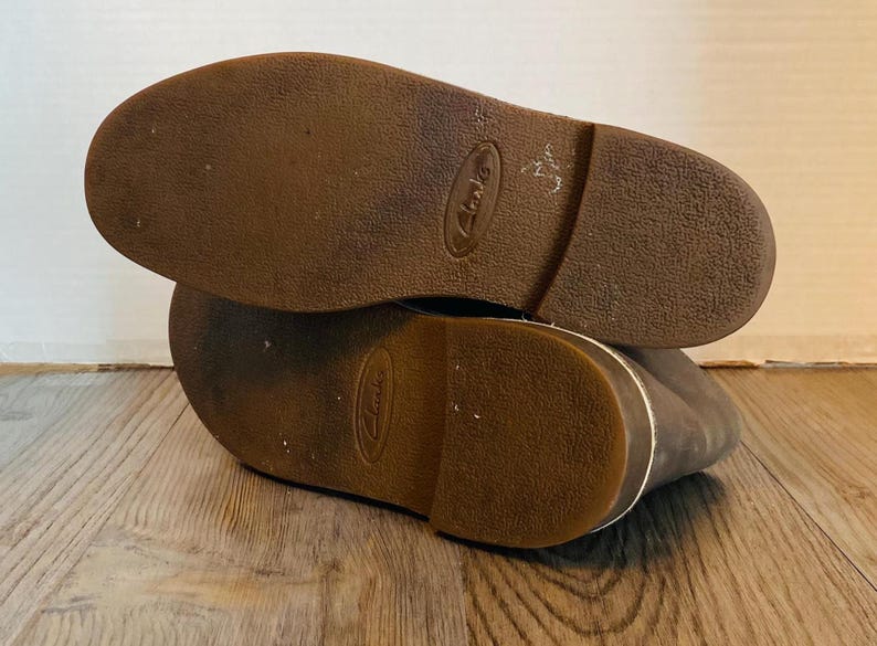 May include: Close-up of the brown leather soles of a pair of brown leather shoes. The shoes have a white stitching detail around the edges of the soles. The brand name "Clarks" is visible on the soles.