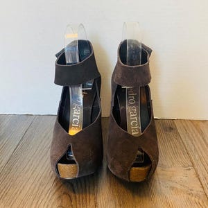 May include: A pair of brown suede peep-toe platform heels. The heels have a cork platform and a strap that wraps around the ankle. The brand name "pedro garcia" is visible on the insole. The shoes are displayed on a wooden surface.