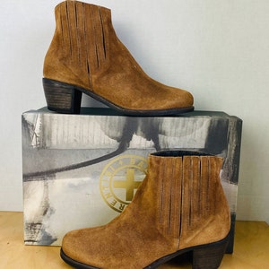 May include: A pair of brown suede ankle boots with a stacked heel. The boots have a zipper on the inside of the ankle and a decorative stitching detail on the back of the boot.