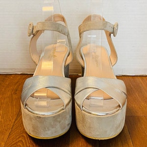 May include: A pair of beige platform sandals with silver straps and buckles. The sandals have a thick platform sole and a high heel.