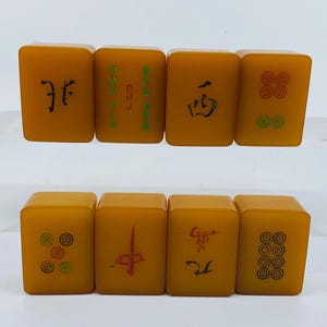 Op de afbeelding: Acht vintage Mahjong-tegels in een warme amberkleur. Elke tegel heeft unieke symbolen en karakters in zwart en groen. De tegels zijn gerangschikt in twee rijen van vier, wat de verscheidenheid aan ontwerpen laat zien.