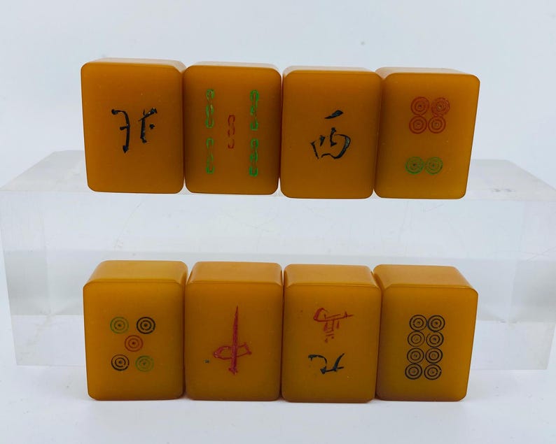 Op de afbeelding: Acht vintage Mahjong-tegels in een warme amberkleur. Elke tegel heeft unieke symbolen en karakters in zwart en groen. De tegels zijn gerangschikt in twee rijen van vier, wat de verscheidenheid aan ontwerpen laat zien.