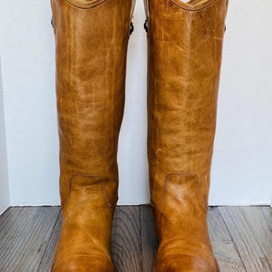 May include: A pair of tall, light brown leather boots. The boots have a classic design with a slightly distressed appearance. They feature a rounded toe, a mid-calf height, and a subtle heel. The leather has a warm, golden hue, and the boots appear well-worn.