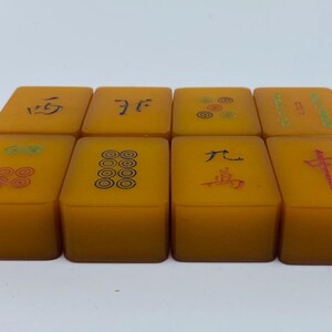 Op de afbeelding: Acht amberkleurige Mahjong-tegels met zwarte en rode symbolen. De tegels zijn gerangschikt in twee rijen van vier, elk met unieke karakters en ontwerpen. De tegels zijn rechthoekig en hebben een enigszins doorschijnende uitstraling.