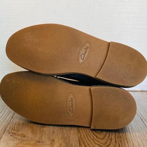 May include: A pair of Clarks brand shoes, soles up, showing the brown rubber soles. The soles have a textured surface and the brand name is embossed on each sole. The shoes are on a wooden surface.