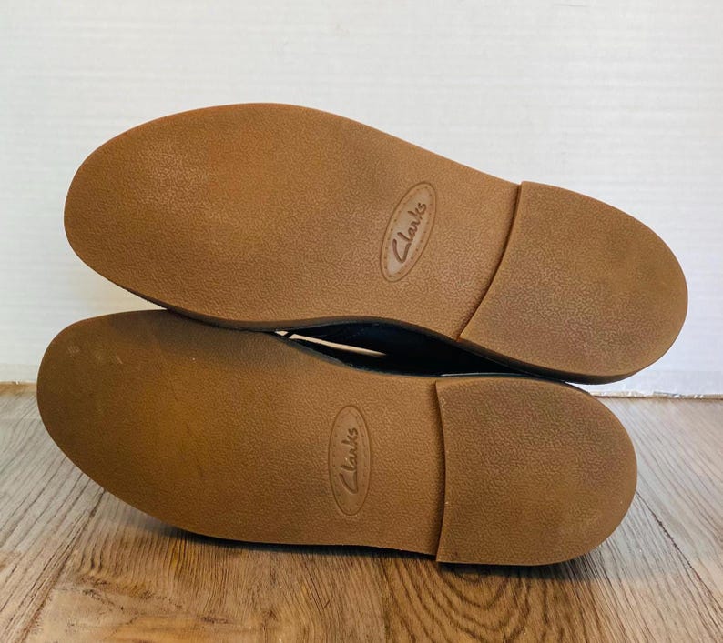 May include: A pair of Clarks brand shoes, soles up, showing the brown rubber soles. The soles have a textured surface and the brand name is embossed on each sole. The shoes are on a wooden surface.