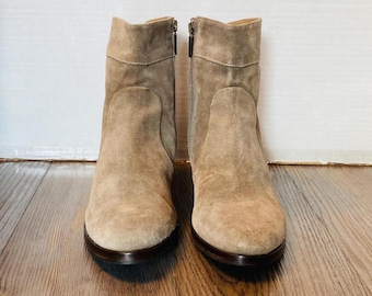 May include: A pair of beige suede ankle boots with side zippers. The boots have a rounded toe and a low heel. The boots are displayed on a wooden surface against a white background. The boots are a neutral color and would complement many outfits.