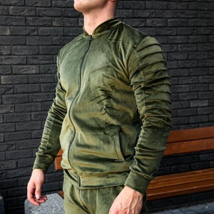 May include: A green velvet tracksuit jacket with a zipper closure and ribbed cuffs. The jacket has a stand-up collar and a pocket on the left side.