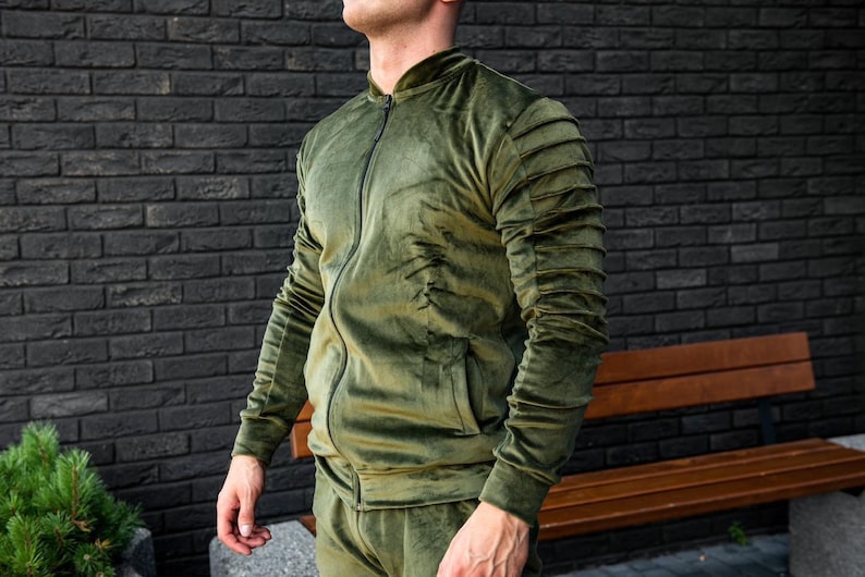 May include: A green velvet tracksuit jacket with a zipper closure and ribbed cuffs. The jacket has a stand-up collar and a pocket on the left side.