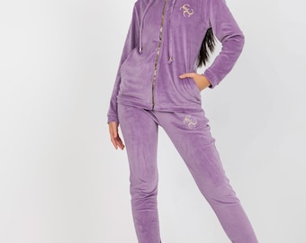 Könnte beinhalten: Ein lilafarbener Trainingsanzug aus Velours mit goldenem Logo. Die Jacke hat eine Kapuze und einen Reißverschluss. Die Hose hat einen Bund mit Kordelzug.