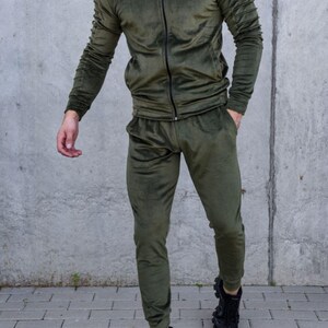 May include: A green velour tracksuit with a zippered jacket and matching pants. The jacket has ribbed detailing on the sleeves. The model is wearing black sneakers.