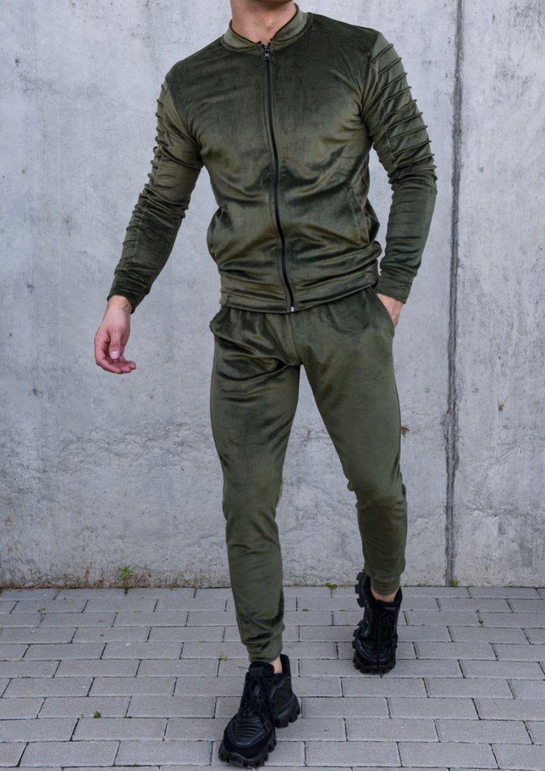 May include: A green velour tracksuit with a zippered jacket and matching pants. The jacket has ribbed detailing on the sleeves. The model is wearing black sneakers.